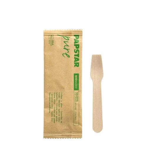 Ein PAPSTAR Eislöffel Holz "pure" 9,4 cm, einzeln verpackt in Papier mit grünem "PAPSTAR pure BIO-BASED"-Aufdruck liegt neben seiner braunen Verpackung von PAPSTAR GmbH auf weißem Hintergrund.